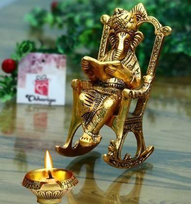 Golden Lord Ganesha statue sitting on a rocking chair reading Ramayan, placed on a polished surface with a lit diya and decorative background.