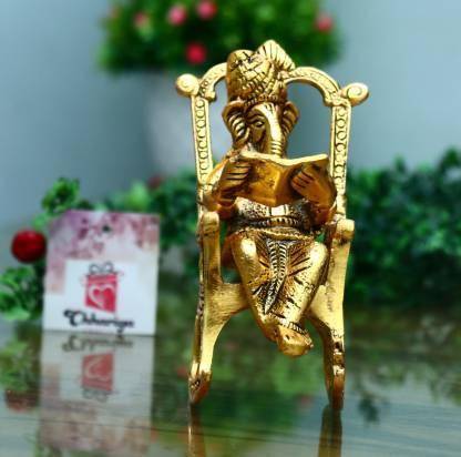 Front view of the Golden Lord Ganesha statue on a rocking chair reading Ramayan, with a blurred green and red decorative background.